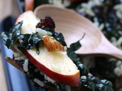 close up salad shot featuring a wooden spoon, almond chuncks, cranberries, kale, and fresh pazazz apples.