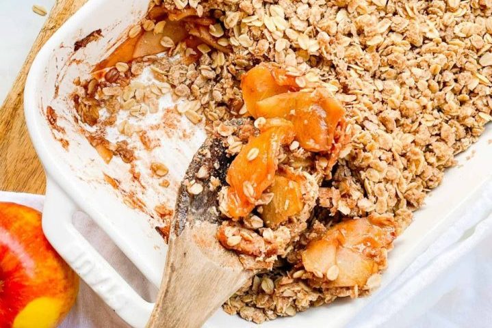 wooden spoon digging into white dish of Pazazz apple crisp with oat topping. All of this sits on a wooden countertop.