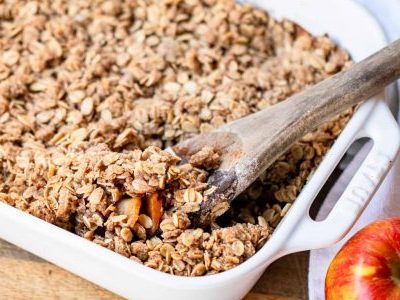 white baking dish filled with Pazazz apple crisp. A wooden spoon is digging into the right side of the crisp. A Pazazz apple sits to the right of the dish. All of this is positioned on a wooden countertop.