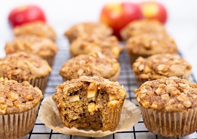 apple muffins sitting in four rows on a metal cooling rack. At the back of the photo three apples are blurred.