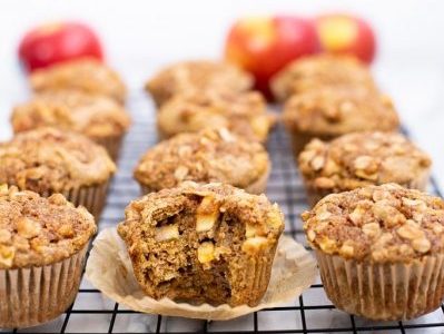 apple muffins sitting in four rows on a metal cooling rack. At the back of the photo three apples are blurred.
