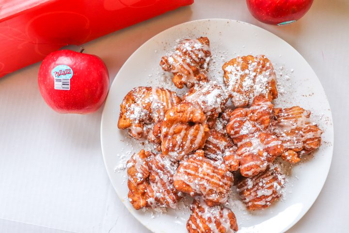 Pile of apple fritters situated on a white plate. They are drizzled with sugar glaze and a sprinkling of powdered sugar. A Pazazz apple sits to the left of the plate and to the upper right. A red apple box sits at the top of the image. All of this is set against a white background.