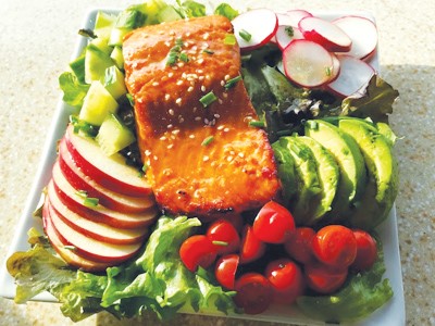 large piece of glazed salmon positioned atop a plate overflowing with fresh vegetables and slices of Pazazz apple. this all sites on a plane colored countertop