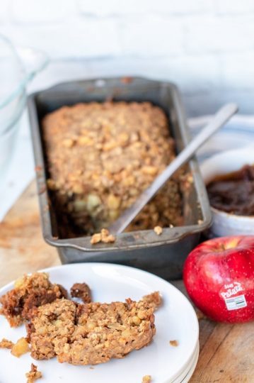 A tin of Oat apple bread sits behind a plate with a slice of bread on it and a bright red Pazazz apple.